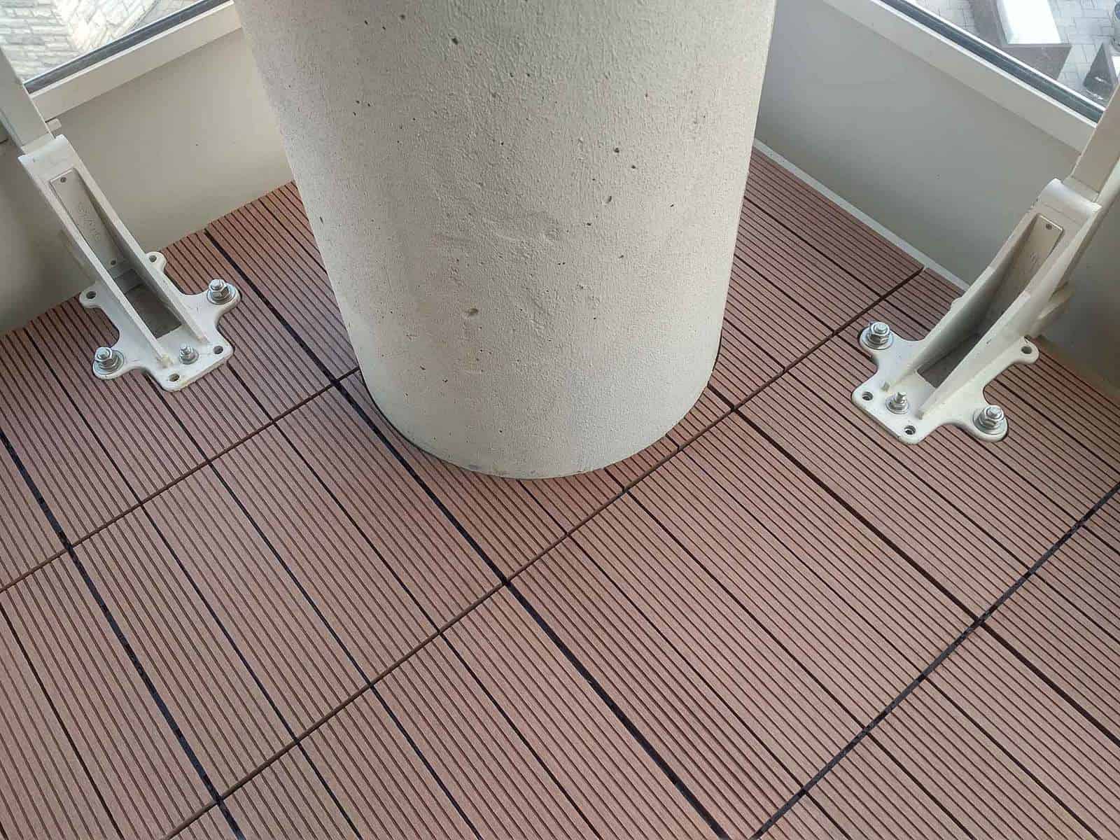 Teak, 4-slat, ridged WPC tile seen on balcony. Measures 1 by 1 feet. Complex cuts were needed to conform to round column and railing posts (footings). Skyscapes green and blue logo at top-left of image.